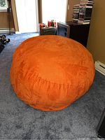 Full view of a large orange bean bag chair on carpeted floor in a basement room with various items in background.