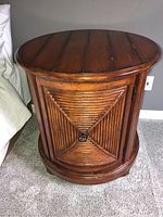 Photo shows front view of round wooden end table with cabinet door featuring a decorative triangular ribbed pattern and metal ring pull handle. The wooden top shows wear.