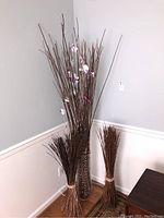Full view of tall clear plexiglass vase filled with clustered brown faux branches with some faux flowers and two tied bundles of plain faux branches on either side.