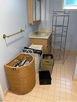 Photo showing large round wicker laundry hamper, white woven rectangular hamper, assorted hangers, small storage boxes, metal 4-tier shelf rack, bathroom vanity and towel rack in basement room.