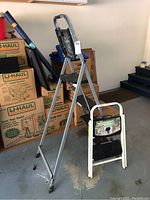 Folding metal painters ladder with four black steps and top platform with paint stains next to a white framed folding step stool with two black steps, all showing wear.