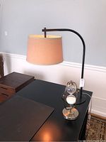 Metal desk lamp with black curved arm and beige fabric drum shade alongside brass-colored hourglass on a black table.
