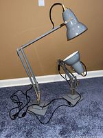 Two gray adjustable arm desk lamps with brass-colored springs and gray bases on carpeted floor against brown wall. One lamp is taller than the other due to arm position.