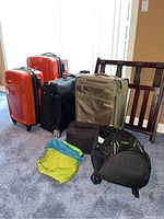 Four pieces of luggage including two orange Samsonite hard shell suitcases, one black soft shell suitcase, one olive green suitcase, black wheeled backpack, green and blue storage tote, and wooden luggage rack on carpet.