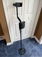 Full length photo of Bounty Hunter Tracker IV metal detector showing shaft, armrest, control box, and search coil on blue carpet in front of white door.