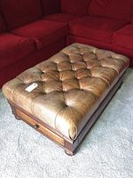 Brown leather tufted cushion ottoman with wood base and drawers placed in front of red sectional sofa