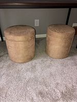 Photo of two matching tan microfiber ottomans placed on carpeted floor under table, showing their cylindrical shape and size