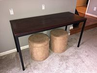 Full view of the sofa table showing the dark wood top and black metal legs, with two cylindrical beige fabric stools underneath.