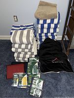 Overall lot view showing towels stacked in blue/white and gray/white stripes, brown throw, black Santa Fe Opera poncho, and packaged rain ponchos on carpet.