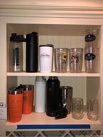 Photo showing two shelves with assorted travel water bottles and plastic cups including CamelBak bottles and Gold Canyon Candles branded tumblers.