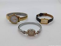Three watches displayed together: white gold watch at front center, Timex Indiglo on left, rectangular watch with leather strap on right