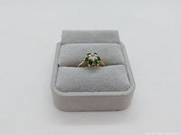Front view of ring in gray velvet display box showing cluster of diamond and emerald stones