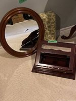 Photo showing the round solid cherry framed wall mirror leaning on a floral patterned box, and the Bombay Company solid cherry tabletop book stand with wavy front slat in front of it, placed on beige carpet.
