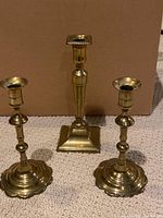 Front view of three brass candlesticks showing the pair with scalloped bases and the taller single with square base.