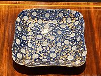 Top-front view of square English porcelain dish with blue, white chrysanthemum and gilt floral pattern on a wooden surface.