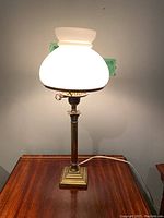 Full view of lit antique desk lamp with milk glass shade on a wood table, showing overall appearance and lighting effect.