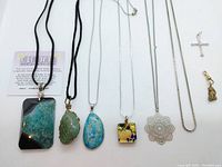 Photo of the full lot showing seven necklaces with various pendants including a large stone pendant, wire-wrapped green stone, blue stone, square Murano glass, filigree metal flower, silver cross, and gold-tone nugget charm