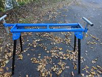 Photo of a blue metal chop saw stand with black legs set on outdoor ground covered with leaves.