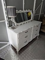 White painted dresser with six drawers, ornate black metal handles, and scalloped drawer fronts, standing on curved legs. Lamp on dresser, metal floor beneath.