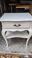 Full view of white wooden nightstand with one drawer and lower shelf with curved legs and scalloped edges.
