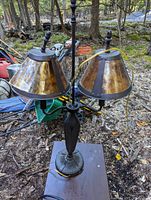 Photo of a double-light table lamp with metal base and brown tinted shades, set outdoors on the ground surrounded by trees and garden equipment.