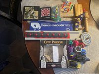 Photo showing assortment of small kids games and toys spread on table, including puzzles, tops, metal cars, cribbage board, Think-It-Thru-Tiles box, and wooden figurines.