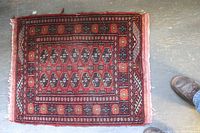 Full view of small rectangular Oriental rug with detailed geometric and floral patterns in red, black, and orange, laid on grey floor with visible foot on side for scale