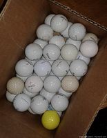 Photo of golf balls in a cardboard box showing multiple white Titleist golf balls and one yellow golf ball, illustrating condition and quantity.