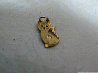 Close-up image of a small gold kitten charm lying on a light patterned surface, showing the textured body, cut-out eyes, and curled tail design.