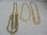 Photo shows three bone necklaces arranged flat on a light patterned fabric background: one multi-strand with small beads, one with medium beads in a single strand, and one with large oval beads