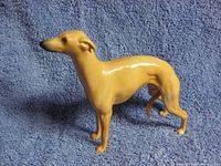 Side view of tan ceramic whippet dog figurine on blue towel background, showing slender form and standing pose.