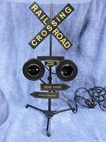 Full view of the vintage railroad crossing light signal on tripod with electrical cord visible, showing crossbuck railroad crossing sign and directional signs reading 'NEW YORK' and 'HOLLYWOOD'.