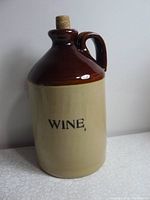 Front view of two-tone ceramic wine jug with cork stopper, beige body with 'WINE' text and brown glazed top and handle.