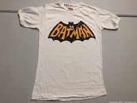 White vintage Batman T-shirt with large black Batman logo and yellow-orange lettering, and Batman face graphic in center.
