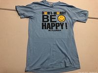 Full front view of the light blue T-shirt showing the Don't Worry Be Happy print with smiley faces.
