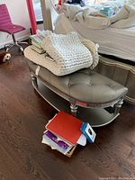 Full view of the tufted bench showing grey leather-like seat with crystal tuft buttons and lower shelf with folded knit blanket and books on and around bench.