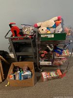 Two metal rolling carts filled with toys, games, puzzles, and other assorted items with additional boxes on the floor