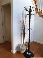 Full view of hardwood coat rack next to metal standing vase with faux flowers and two smaller flower arrangements on floor.