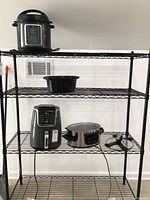 Wide shot showing three appliances on metal shelving: top shelf has Mealthy MultiPot pressure cooker, second shelf has a large black pot insert, third shelf has Ninja Air Fryer XL and Crockpot pressure cooker.