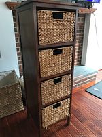 Front view of tall wooden cabinet with four woven baskets installed as drawers, placed on wood floor in front of brick fireplace. Cabinet has visible scratches on wood surface.