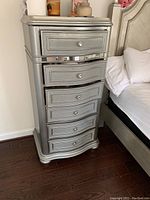Full view of one silver lingerie cabinet showing six drawers, crystal knobs, and curved base on dark hardwood floor next to bed.