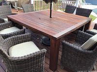 Photo of wooden square patio table with a slatted top, central hole with umbrella pole, showing surface wear and weathering. Surrounded by wicker chairs (not included).