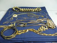 Top view of assorted gold toned costume jewelry laid on blue cloth showing bead necklace, various chain link necklaces and bracelets, smooth bangles, and a heart pendant.
