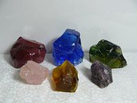 Six chunks of slag glass and minerals arranged on white surface, showing colors and textures.