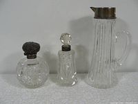 All three cut glass vanity pieces arranged in a row on white surface against plain background. Shows shape and details of pieces: two perfume bottles and a pitcher.