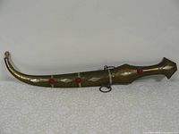 Full view of curved eastern ceremonial dagger with ornately decorated metal sheath and red bead accents lying flat on a surface.