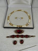 Formal costume jewelry set in display case including cream and gold-tone necklace with matching earrings, a red gemstone brooch, a red gemstone bracelet, and red gemstone earrings.