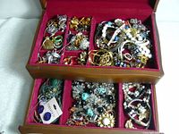 Photo of upper compartments of the jewelry box showing gold and silver-tone rings, earrings, brooches, and pendants in multiple small sections with varied designs and materials including enamel and beaded accents.