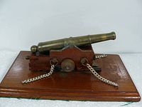 Side angle showing brass cannon mounted on wooden carriage and base, with two brass chains extending from carriage sides.
