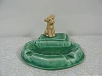 Whole view of the vintage green ceramic dish topped with a beige puppy figurine on raised cushion.